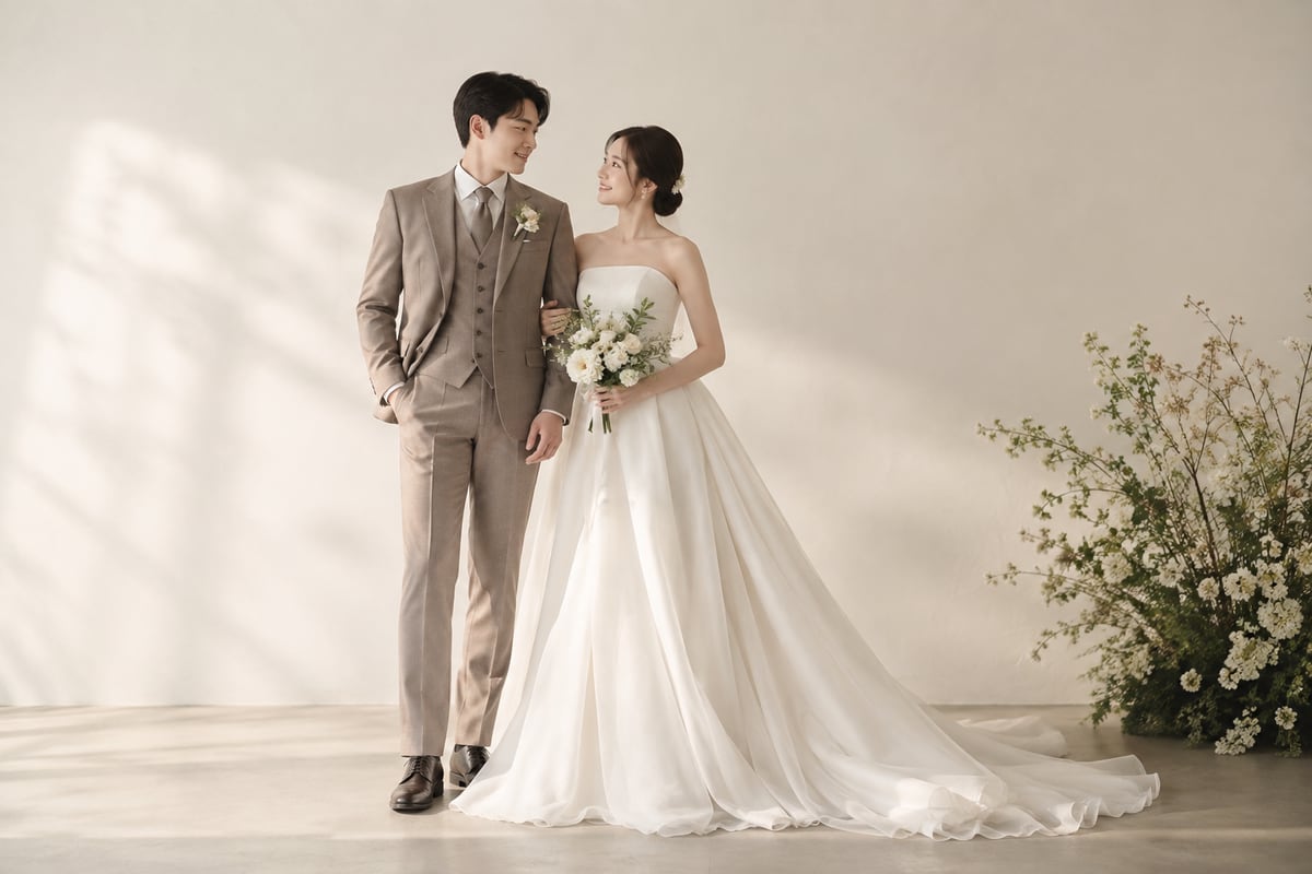 밝은 아이보리 배경 앞에서 아이보리 드레스와 회갈색 예복을 입은 신랑 신부